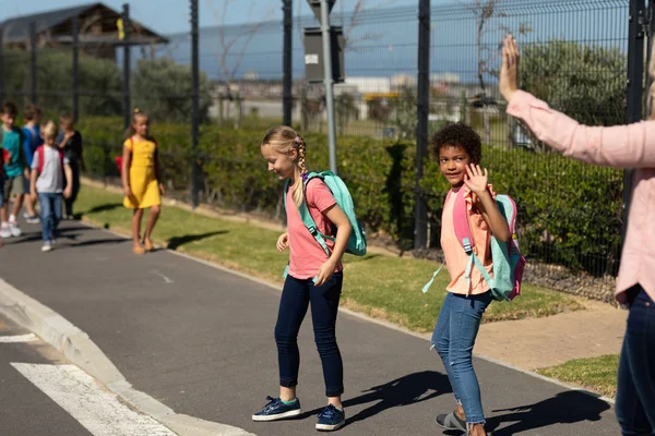アフリカ系アメリカ人に手を振っている白人女性教師と 晴れた日に小学校に行く途中の歩行者交差点で道路を横断するのを待っている白人女の子の側面図 — ストック写真