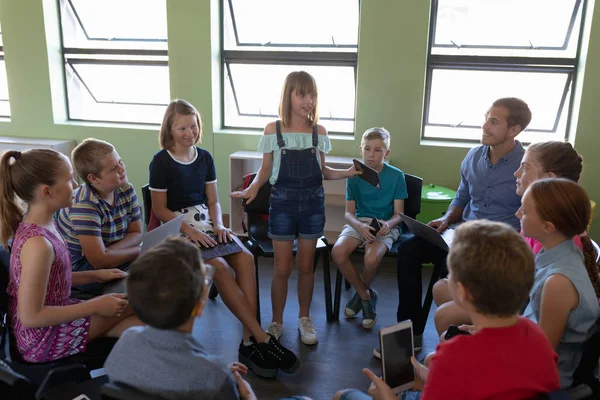 Front view of a diverse group of elementary school kids sitting on chairs in a circle and interacting during a lesson, one Caucasian girl standing and talking while her classmates and male Caucasian teacher sit and listen