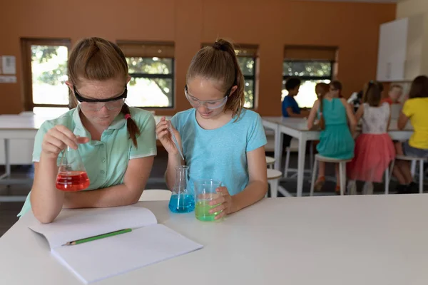 小学生の理科の実験をしながら 安全眼鏡をかけカラフルな液体のフラスコを使って机に座っている2人の白人女子学生の正面図 — ストック写真