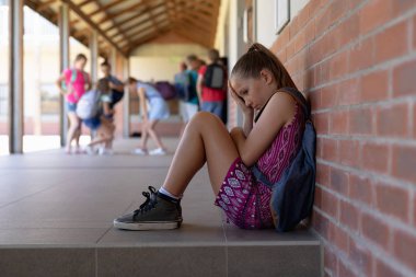 Side view of a Caucasian schoolgirl wearing a purple dress and a rucksack sitting on the ground against a wall alone in the schoolyard at elementary school looking sad, with other schoolchildren standing together in the background clipart