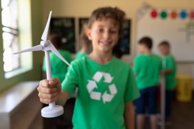Front view of a Caucasian schoolboy with short dark hair wearing a green t shirt with a white recycling logo on it, holding miniature wind turbine and looking to camera smiling in an elementary school classroom clipart