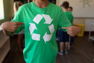 Front view mid section of schoolgirl wearing a green t shirt with a white recycling logo on it and pointing at it, in an elementary school classroom, with her classmates standing in the background clipart
