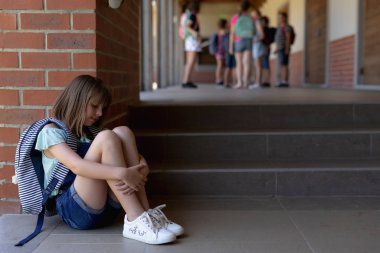 Side view of a Caucasian schoolgirl wearing denim shorts, sports shoes and a rucksack sitting on the ground alone in the schoolyard at elementary school looking sad, with other schoolchildren standing together in the background clipart