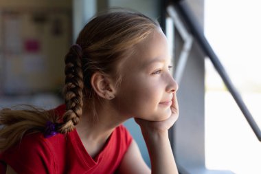 Portrait of a Caucasian schoolgirl with blonde hair in plaits wearing a red t shirt, sitting at a desk resting his chin on her hand and looking out of an open window in thought during a lesson in an elementary school classroom clipart