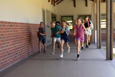 Front view of a diverse group of schoolchildren wearing shorts, skirts and rucksacks running in an outdoor corridor at elementary school clipart