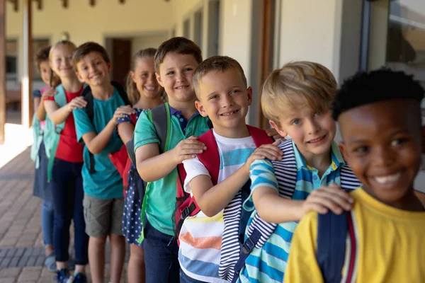 Front view of a diverse group of schoolchildren walking single file with hands on each others shoulders along an outside corridor between classrooms at an elementary school, looking to camera and smiling