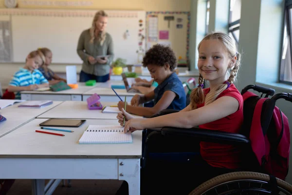 机の上の車椅子に座って カメラに目を向け 机の横の小学校の教室で働いている子どもたちと笑顔で座っている白人の女子学生の姿 — ストック写真