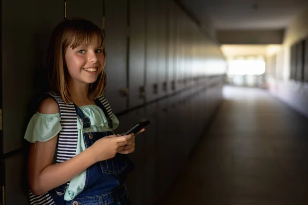 スマホを使って廊下のロッカーにもたれかかるリュックサック姿の白人女子高生の横からの眺めや 小学生の時のカメラへの笑顔 — ストック写真