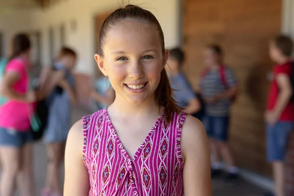 Portrait of a Caucasian schoolgirl wearing a purple top standing in the schoolyard at elementary school smiling to camera, with other schoolchildren standing in the background