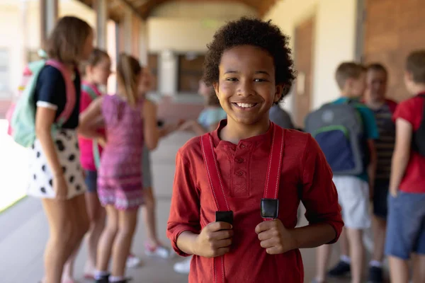 アフリカ系アメリカ人の少年がカメラに微笑む小学校の校庭に立ったリュックサックを背景に他の子供たちが立っている姿 — ストック写真
