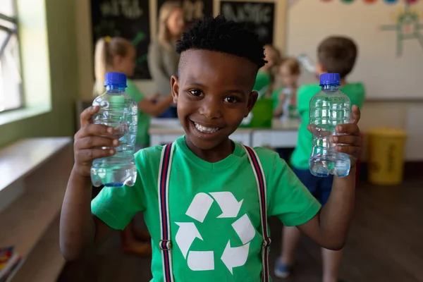 白いリサイクルのロゴが入った緑色のTシャツを着たアフリカ系アメリカ人の少年の肖像画 2本のペットボトルを持ち カメラを探して笑顔で 小学校の教室で — ストック写真