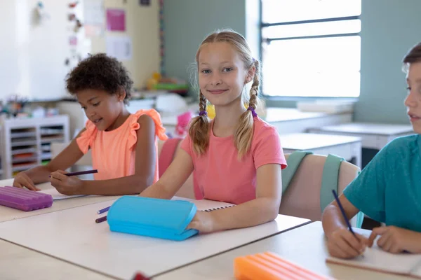 在小学课堂上 一个头戴粉色T恤 头戴金黄色斑斑的白人女学生坐在课桌前 微笑着对着摄像机拍照的正面景象 — 图库照片