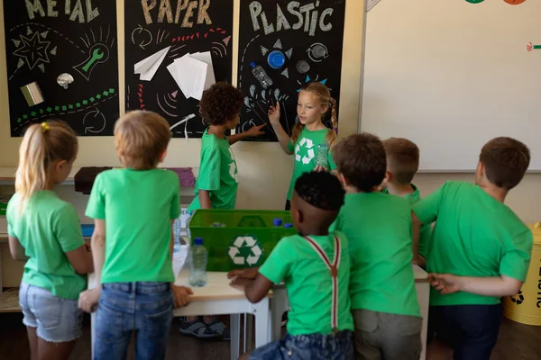一群身穿绿色T恤衫的学童站在回收箱周围 听着一位白人和一位非裔美国女学生站在那里与他们交谈 并指着一张可回收材料的海报 — 图库照片