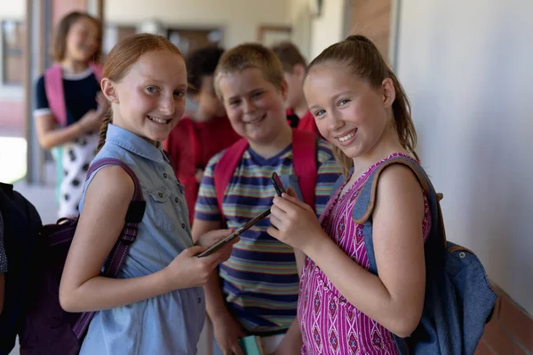 2人の白人女子校生と1人の白人男子校生が 小学生の時にバックパックを着てカメラに向かって笑顔で立ち 1人の女の子がタブレットコンピュータを持っている — ストック写真