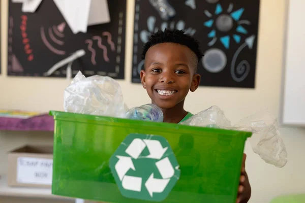 一个短头发的非洲裔美国学生的画像 他拿着一个绿色的箱子 上面有一个白色的回收标识 他期待着在小学教室里微笑着拍照 — 图库照片