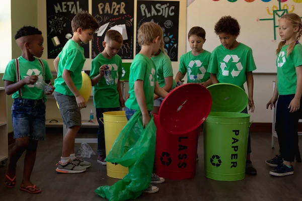 不同类型的学童穿着绿色T恤 上面有白色环保回收标志 在小学课堂上拿着色标回收箱和回收袋的侧视图 — 图库照片