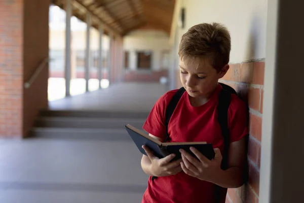 一个身穿背包和红色T恤的白人学童靠着墙在室外走廊边看书 在小学里的镜头映入眼帘 — 图库照片