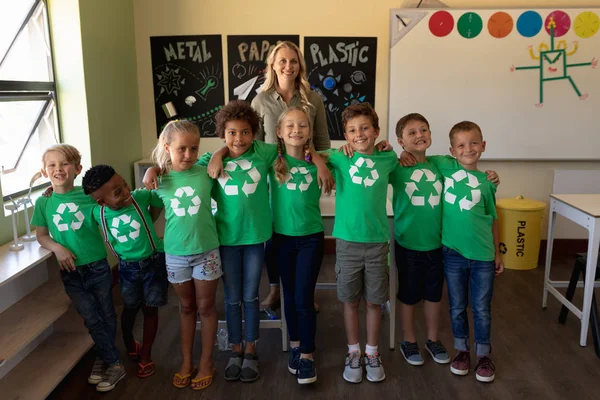 ブロンドの髪をした白人女性教師の肖像画と 白いリサイクルロゴが入ったグリーンのTシャツを着た8人の生徒の多様なグループが小学校の教室に立っている — ストック写真