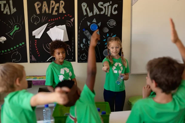 小学校の教室でリサイクル可能な教材についてのポスターの前に立つ白人とアフリカ系アメリカ人の女子学生に質問するために手を挙げて緑のTシャツを着た多様な子供たちの後ろのビュー — ストック写真