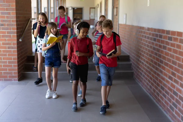 小学校の読書本の外廊下を歩いたり ヘッドフォンをしたり 電話をしたり 話をしたりする多様な生徒たちの前の風景 — ストック写真