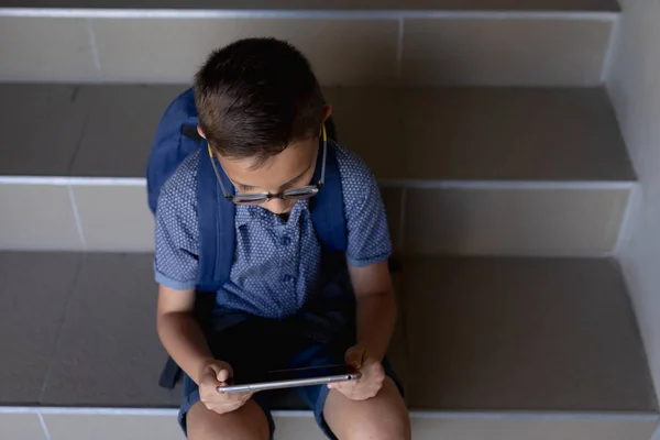 タブレットコンピュータを使用して 小学校の階段に座ってメガネ ショートパンツ リュックサックを身に着けている白人の学童の高角度ビュー — ストック写真