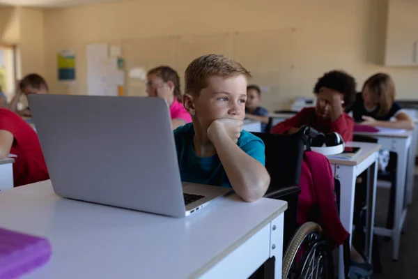 小学校の教室でノートパソコンを使って机の上に座って手にもたれかかって思考を逸らす白人の少年の正面図 — ストック写真
