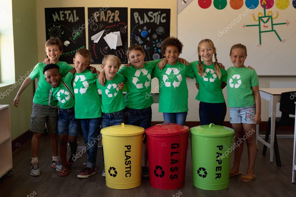 Portrait of a diverse group of schoolchildren wearing green t shirts with a white recycling logo on them, standing behind color coded recycling bins in an elementary school classroom with arms around each other, smiling to camera