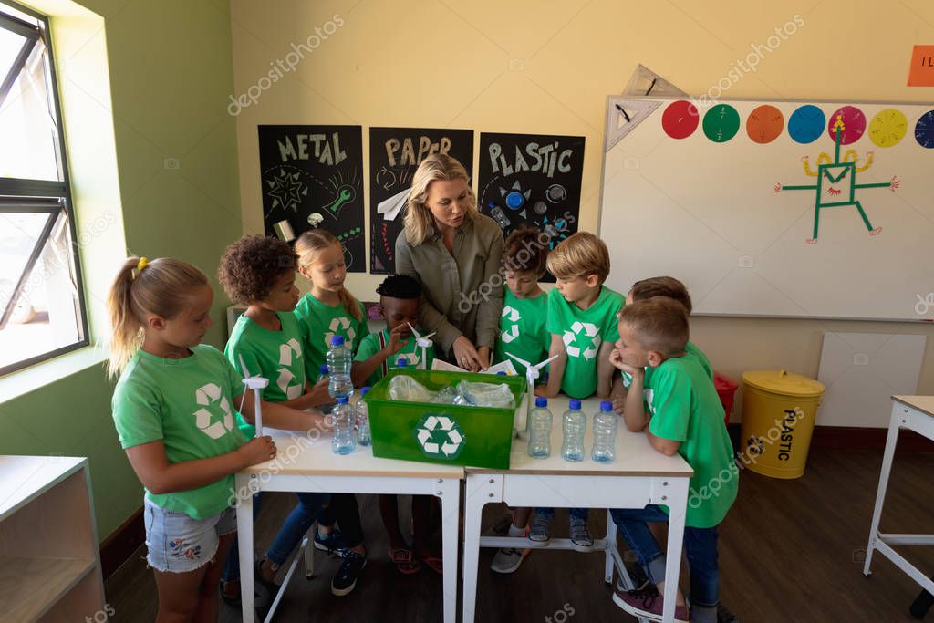 Front view of a Caucasian female school teacher with long blonde hair and a diverse group of schoolchildren wearing green t shirts with a white recycling logo on them standing around a recycling box on a table