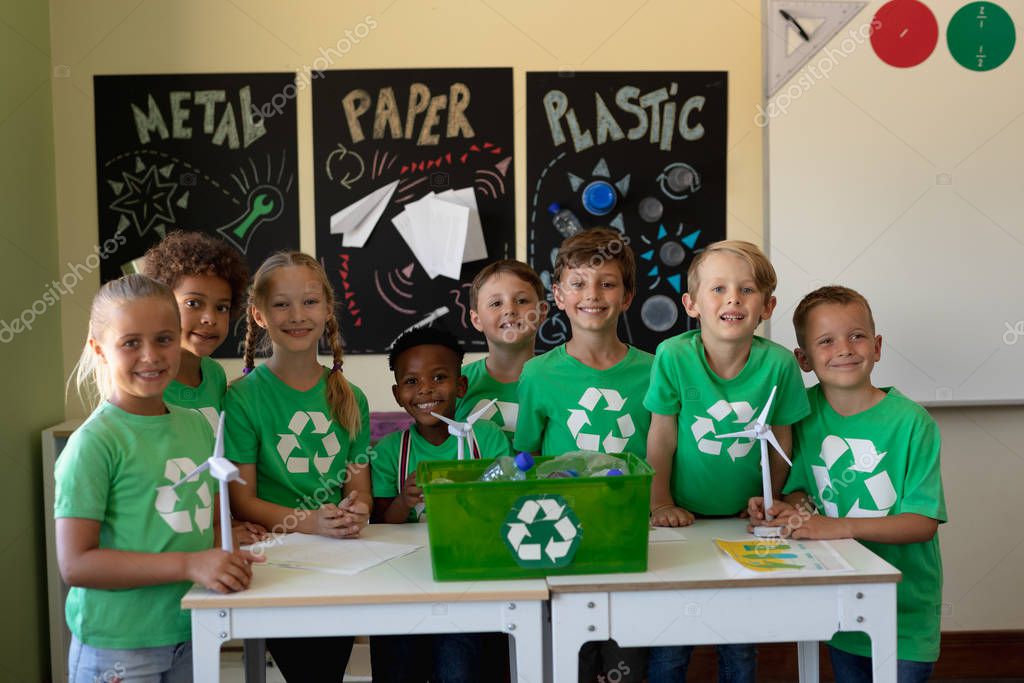 Portrait of a diverse group of eight schoolchildren wearing green t shirts with a white recycling logo on them standing around a recycling box on a table and holding three miniature wind turbines in front of posters about recyclable materials