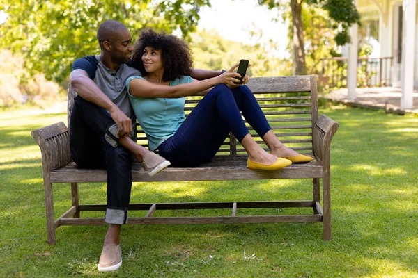 一对混血儿坐在花园里的长椅上 面带微笑 手里拿着一部手机 眼前的景象 — 图库照片