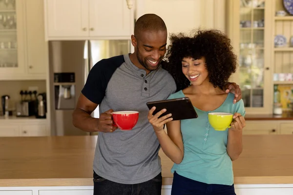 一对混血种族的夫妇站在厨房里 举杯咖啡 看平板电脑的镜头 — 图库照片