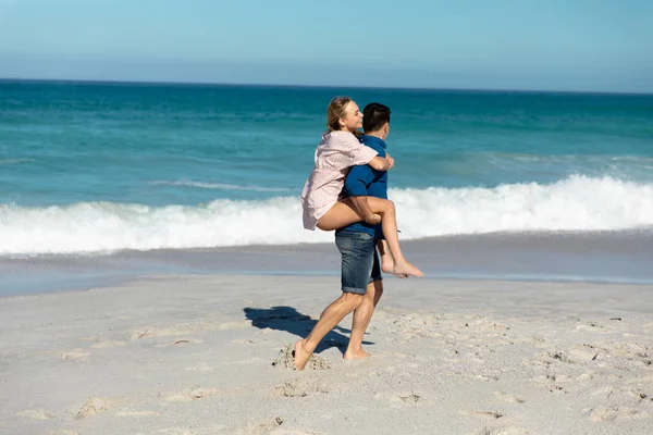 背景に青い空と海のビーチで白人のカップルの側のビュー 砂の上を歩く ピギーバックと笑顔 — ストック写真