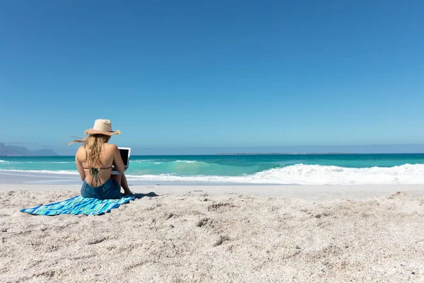 ビーチのタオルの上に座っている白人女性の後部ビューでは 彼女のラップトップを使用して 背景に青い空と海を持つわら帽子を身に着けています — ストック写真