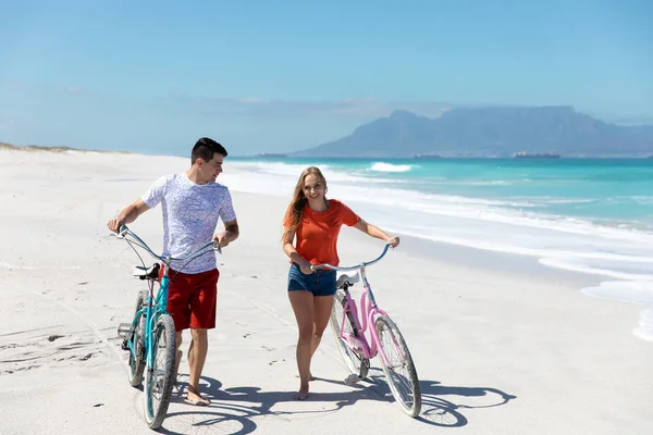 青い空と海を背景にビーチを歩く白人のカップルのフロントビュー 笑顔と彼らの自転車をリード — ストック写真