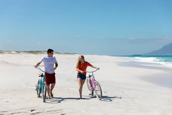 青い空と海を背景にビーチを歩く白人のカップルのフロントビュー 笑顔と彼らの自転車をリード — ストック写真