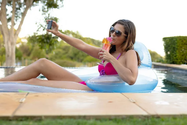 ビーチウェアを着た白人女性と 晴れた日にプールで日光浴をするインフレータブルプールのラウンジャーに座ってサングラスをかけ カクテルを飲み スマートフォンで自撮り — ストック写真
