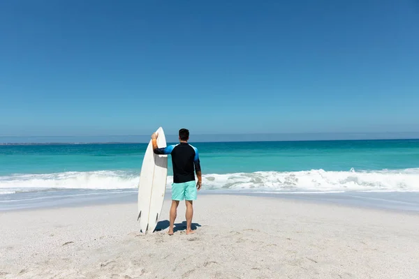 背景に青い空と海を背景に海岸に立っている白人男性の後部ビュー サーフボードを保持し 離れて見て — ストック写真