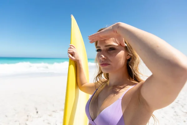 青い空と海を背に海岸に立つ白人女性の側面図サーフボードを持って遠くを見る — ストック写真
