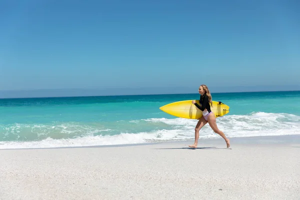 青い空と海を背に海岸を歩く白人女性のサイドビュー サーフボードを保持 — ストック写真