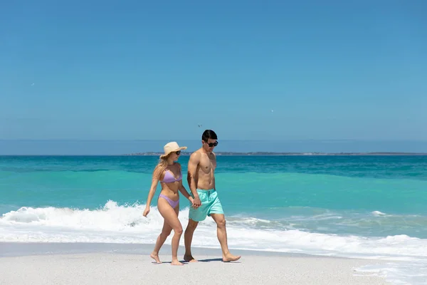 青い空と海のビーチを背景に 手を取り合って笑顔で歩く白人のカップルの側面図 — ストック写真