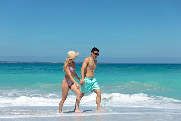 一对白种人夫妇手牵手 面带微笑地在沙滩上走着 背景是蓝天蓝海 — 图库照片