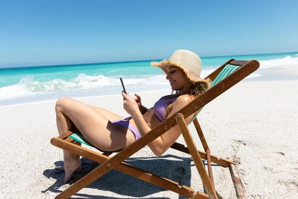 青い空と海を背景にビーチで机の上に座って笑顔と彼女の携帯電話を使用して白人女性の側面図 — ストック写真