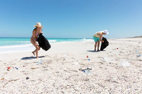 青い空と海を背景にしたビーチでの白人夫婦のフロントビュー ゴミを拾い ゴミ袋に入れる — ストック写真