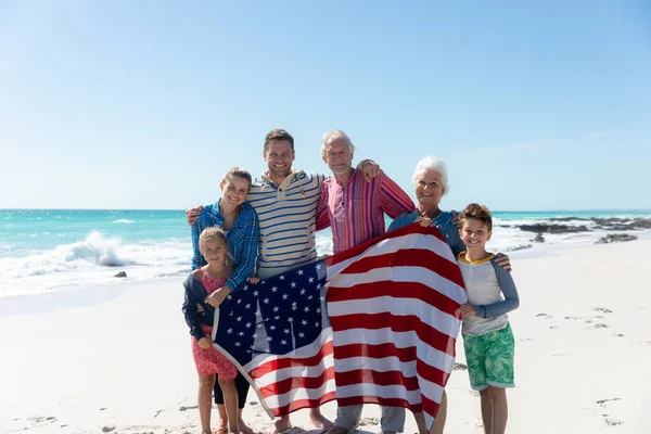 背景には青い空と海が広がるビーチに立つ多世代の白人家族のフロントビュー アメリカ国旗の掲揚 カメラへの抱きしめと笑顔 — ストック写真