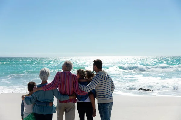 一个多代白人家庭站在海滩上的后视镜 背景是蓝天和大海 拥抱和远眺 — 图库照片