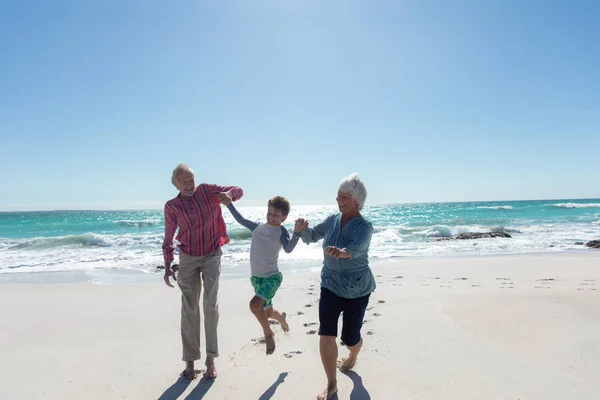 祖父母と一緒に 青い空と海を背景にビーチを歩き 手を取り合い 楽しい時間を過ごしている白人の少年の側面図 — ストック写真