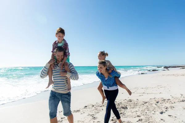 背景是蓝天蓝海的海滩上 一个白人家庭的正面景象 他们一边走路 一边骑着小车 一边笑着 — 图库照片