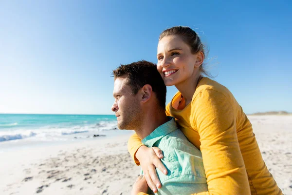 背景に青い空と海のビーチに立っている白人のカップルの側面図 ピギーバック 笑顔と離れて見て — ストック写真