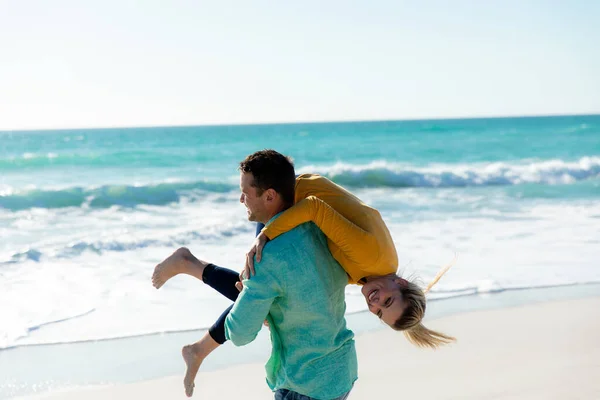 一个白种人背着他的白种人女友走在沙滩上 背景是蓝天蓝海 面带微笑 — 图库照片
