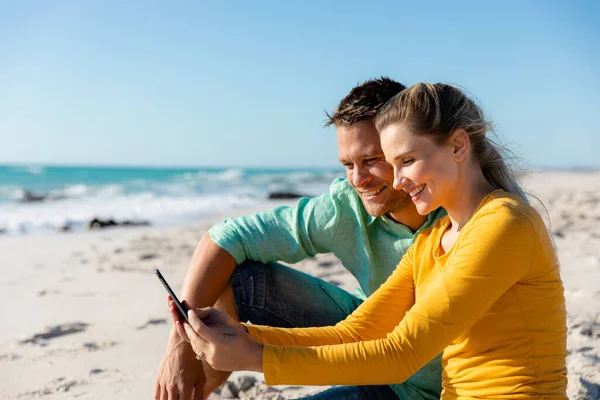 一对白种人夫妇斜倚在沙滩上 背景是蓝天和大海 他们拥抱并看他们的智能手机 — 图库照片